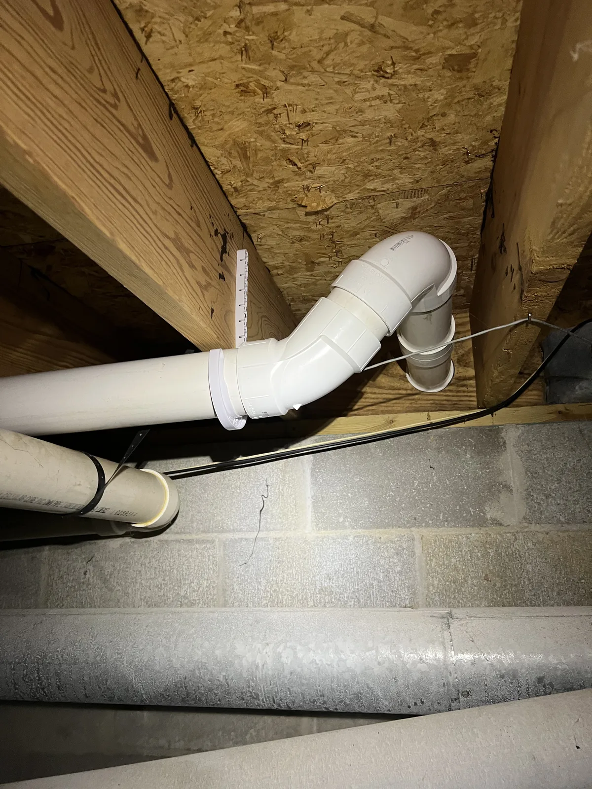 White PVC pipe connection and elbow mounted beneath a wood-framed floor